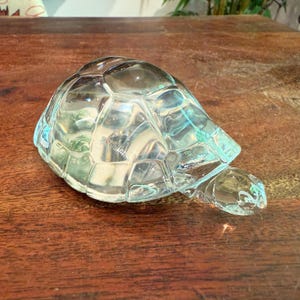 May include: A clear glass turtle ornament with a detailed shell and head. The ornament is transparent, allowing light to pass through, and sits on a wooden surface. The turtle is a decorative item.
