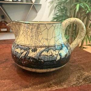 May include: A ceramic pitcher featuring a cream-colored base with a prominent dark crackle glaze finish. The lower section is decorated with a blue and black hand-painted scene depicting two sailing ships on water, complete with a curved handle and spout.