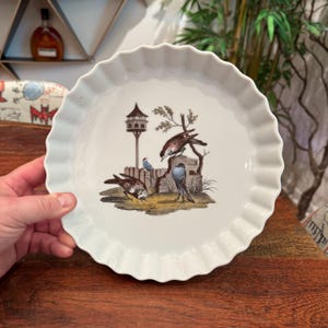 Puede incluir: Plato de tarta de cerámica blanca con borde ondulado. El plato presenta una ilustración detallada de varios pájaros, una casa para pájaros y una pequeña valla. Los pájaros son en tonos marrones, azules y grises.