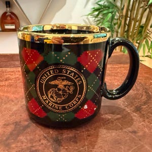 May include: A black ceramic mug featuring a gold rim and a repeating red and green argyle pattern. The center displays a gold circular emblem with the text United States Marine Corps surrounding an eagle, globe, and anchor graphic design.