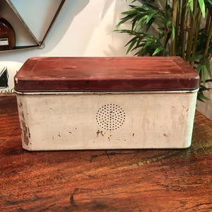 May include: A rectangular, vintage bread box with a white body and a maroon lid. The box has a circular pattern of small holes on the front and shows signs of wear, suggesting its age. The bread box is made of metal.