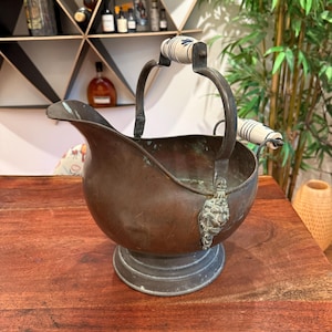 May include: Antique copper coal scuttle with a spout and a handle with ceramic accents. The scuttle has a decorative lion's head detail and a round base. The ceramic handles have a blue floral design.