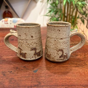 May include: Two speckled beige ceramic mugs with a cylindrical shape and curved handles. Each mug features a horizontal band with a geometric pattern and a series of small, dark brown animal figures etched into the lower section of the exterior.