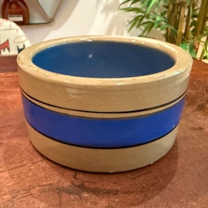 May include: A cylindrical ceramic bowl with a beige exterior and a vibrant blue interior. The outer surface features a wide horizontal blue band encircling the middle, bordered by two thin black lines, creating a simple and functional design.