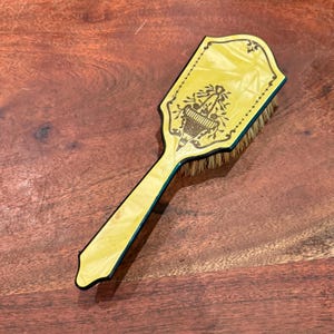 May include: A yellow hairbrush with a black border and a black printed graphic of a floral urn on the back. The handle is long and slender, and the brush features natural-colored bristles extending from the base of the head.