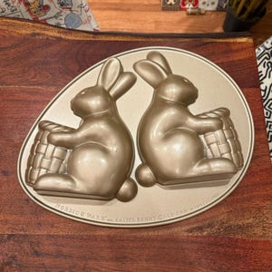 May include: A gold-coloured Easter bunny cake pan with two bunny moulds, each holding a basket. The pan is oval-shaped and has the text "Nordic Ware" and "Easter Bunny Cake Pan" printed on it. The pan holds 2.4 litres.