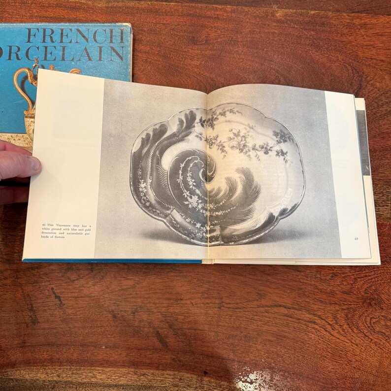 May include: An open book displaying a black and white image of a decorative plate with floral and wave-like patterns. The plate is on a page with text. A blue book with the words "FRENCH PORCELAIN" is visible.