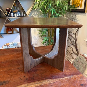 Puede incluir: Una mesa auxiliar cuadrada de madera con una tapa marrón oscuro y una base única de patas cruzadas. La mesa mide aproximadamente 61 cm de ancho y 61 cm de largo, con una altura de unos 51 cm. La veta de la madera es visible.