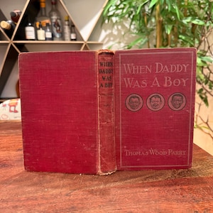 May include: A vintage red hardcover book titled When Daddy Was A Boy by Thomas Wood Parry. The front cover features the title in white text above three circular illustrations of faces. The spine also displays the title in black lettering.