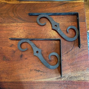 May include: Two black metal shelf brackets with a decorative scroll design. The brackets are L-shaped, with a flat base and a curved, ornate top. The brackets are made of a dark metal and are designed to be mounted on a wall.