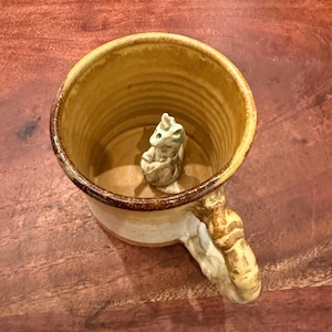 May include: A ceramic mug with a glossy honey-colored interior and a textured, light-colored exterior. Inside the mug, a small, sculpted, light-colored creature figure sits at the base. The mug features a thick, textured handle with a brown rim.