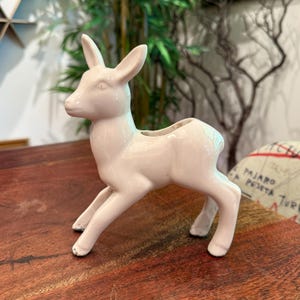 Puede incluir: Figura de ciervo de cerámica rosa claro con la parte trasera hueca, probablemente para sostener pequeñas plantas o flores. El ciervo está de pie con la cabeza ligeramente girada hacia la izquierda. La figura tiene un acabado liso y brillante.