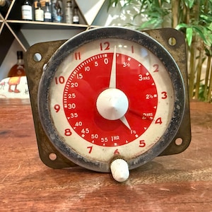 May include: A vintage square-faced industrial timer with a circular dial featuring a red center and white outer ring. The face displays numbers 1 through 12 in red, with smaller minute and hour markings. A white central knob and a small white adjustment knob at the bottom are present. The metal casing shows signs of age and wear, with the text 'FERMURY CONN. U.S.A.' printed at the bottom edge.