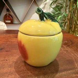 May include: A ceramic apple-shaped jar with a glossy yellow finish and a red blush on one side. The lid features a sculpted green stem and leaf handle. The container has a rounded body and a smooth, reflective surface, designed for storage.