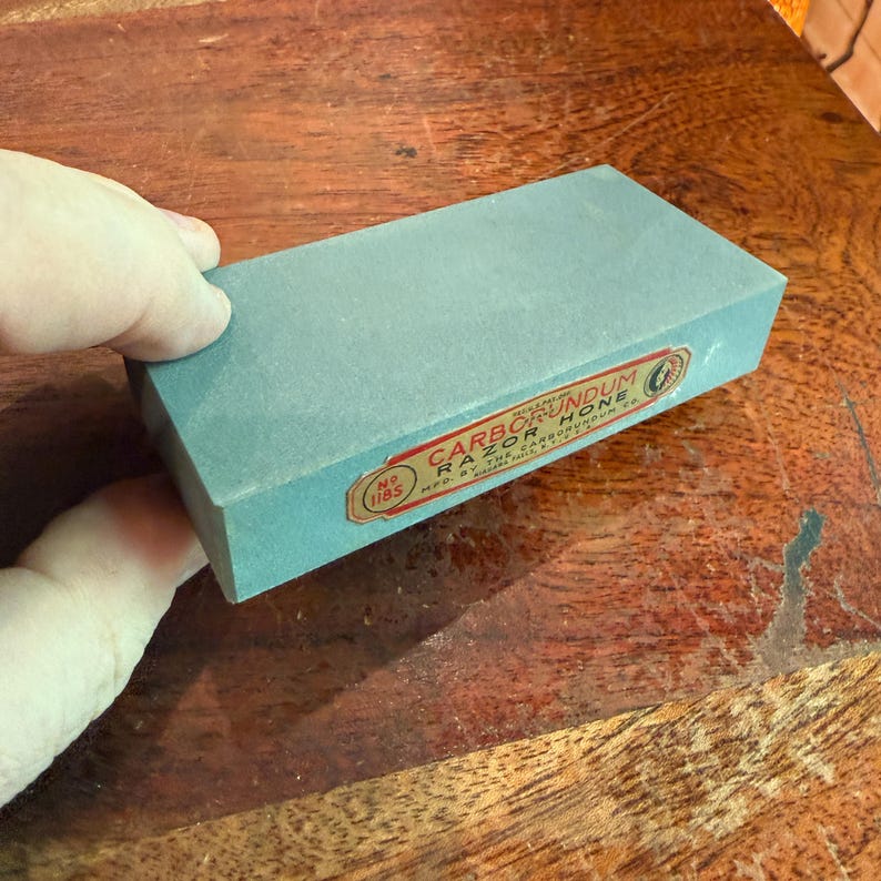 May include: A rectangular, light blue Carborundum Razor Hone sharpening stone. The stone has a label with red and white text. The stone is held by a hand, with the background showing a wooden surface.