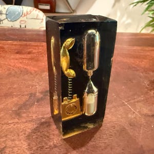 May include: A rectangular black resin block containing a gold-colored miniature rotary telephone and a small glass hourglass filled with white sand. The objects are embedded within the dark, semi-transparent material, creating a clear contrast.