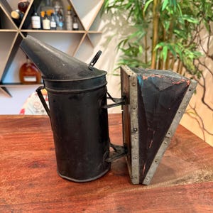 May include: A black metal bee smoker with a bellows. The smoker has a cylindrical body with a conical top and a handle. The bellows are made of wood and leather, with metal trim. The smoker is designed to produce smoke for beekeeping.