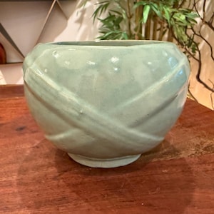 May include: A round, light mint green ceramic bowl featuring a smooth, glossy finish and a decorative pattern of raised, intersecting diagonal bands that wrap around the exterior surface, creating a subtle texture and a rounded, bulbous silhouette.