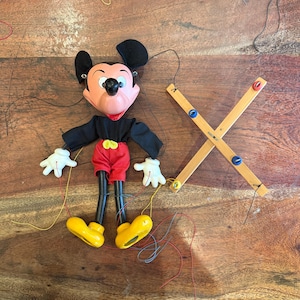 Puede incluir: Una marioneta de Mickey Mouse con una cara pintada, orejas negras y una camisa negra. La muñeca lleva pantalones cortos rojos, zapatos amarillos y guantes blancos. El control de madera tiene forma de X.