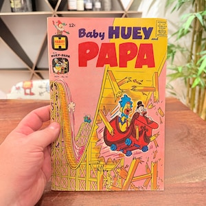May include: A vintage Baby Huey and Papa comic book with a pink and yellow cover featuring a cartoon duck riding a red roller coaster car that is breaking through wooden tracks. The title text reads Baby Huey and Papa with a 12 cent price tag.