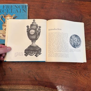 May include: An open book displays a detailed illustration of a decorative clock with a pedestal base. The clock is ornate with a round face and a domed top. A book titled "French Porcelain" is visible in the background.