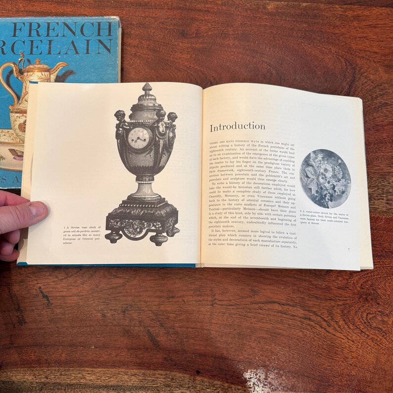 May include: An open book displays a detailed illustration of a decorative clock with a pedestal base. The clock is ornate with a round face and a domed top. A book titled "French Porcelain" is visible in the background.