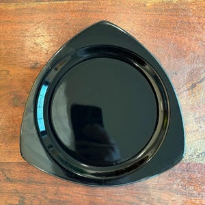 May include: A glossy black plate with a unique triangular shape. The plate features a circular indentation in the center and a smooth, reflective surface. The plate is designed for serving food and adds a modern touch to any table setting.