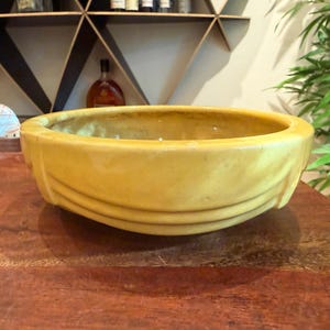 May include: A round, mustard yellow ceramic bowl with a smooth, glossy finish. The exterior features a series of three horizontal, raised, curved bands that wrap around the circumference of the vessel, creating a subtle, layered, and textured appearance.