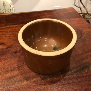 May include: A brown ceramic crock with a light brown rim. The crock has a glazed interior and is slightly weathered.