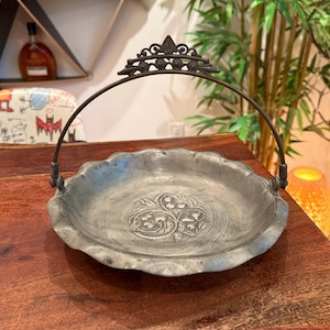 May include: A vintage metal serving dish with a scalloped edge and a decorative handle. The dish has an embossed floral design in the center and a dark, aged finish. The handle is arched and attached to the dish with ornate brackets.