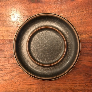 May include: A dark gray ceramic saucer with a smaller saucer nested inside. Both saucers have a brown rim.