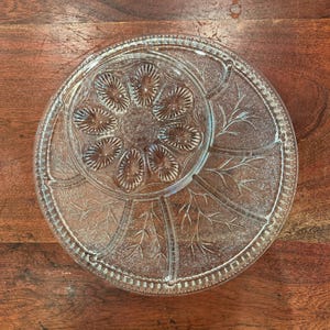 Peut inclure: Présentoir à gâteau en verre transparent avec un motif floral et de feuilles. Le présentoir a une base ronde et un centre légèrement surélevé, parfait pour présenter des gâteaux ou d'autres desserts. Le diamètre est d'environ 30 cm.