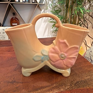 Könnte beinhalten: Eine pfirsichfarbene Keramik-Doppelvase mit einem zentralen Bogengriff und zwei ausgestellten Öffnungen. Die Außenseite ist mit einer erhabenen, hellrosa Blume und kleinen bläulich getönten Blättern verziert und steht auf zwei geschwungenen Füßen.