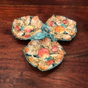 May include: A three-section ceramic serving dish with a turquoise handle. The dish is decorated with a leafy pattern and painted with red, orange, and yellow fruit.