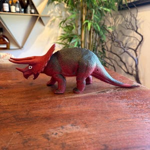 May include: A red and dark green Triceratops toy dinosaur with horns and a frill. The toy is made of plastic and is posed on a wooden surface. The dinosaur has a long tail and is in a walking position.