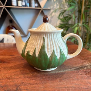 May include: White ceramic teapot with a green and white corn design. The teapot has a brown knob on the lid.