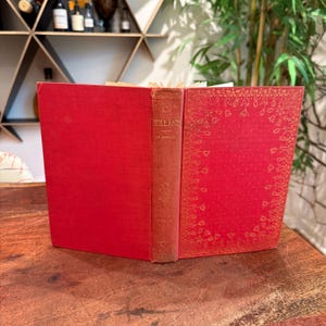 Peut inclure: Livre ancien ouvert avec une couverture et un dos rouges. Le dos porte l'inscription "HOLLAND DE AMICIS" en lettres dorées. Les pages ouvertes ont un fond rouge avec un motif floral doré. Le livre est posé sur une surface en bois.