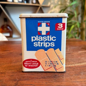 May include: A vintage metal tin of White Cross plastic strips. The tin is white with a blue panel featuring a white cross and the words "plastic strips." It includes three sizes of adhesive bandages: 30-¾ in x 3 in, 15-⅝ in x 1½ in, and 5-1 in x 3 in.