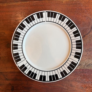May include: White ceramic plate with a black piano key border. The plate has a glossy finish and a circular shape. The piano key design is a repeating pattern around the edge of the plate. The plate is on a dark wood surface.