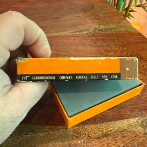 May include: An open orange box with a dark gray rectangular object inside. The box has the text "THE CARBORUNDUM COMPANY, NIAGARA FALLS, NEW YORK" printed on it. The box is held by a hand.