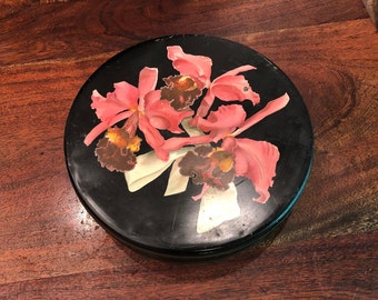 Puede incluir: Una caja redonda de metal negro con un diseño floral. La tapa presenta un grupo de orquídeas rosas con centros marrones y detalles dorados sobre un fondo negro.