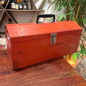 May include: A rectangular red metal toolbox featuring a black plastic handle on top and a silver metal latch closure on the front. The exterior surface shows signs of wear with visible scratches and minor paint chips across the body.