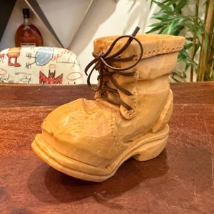 Peut inclure: Une sculpture en bois beige clair en forme de botte de travail avec un bout arrondi, une semelle épaisse et des lacets en cuir marron foncé noués sur le devant. La surface présente des marques de ciseau visibles et un grain de bois naturel.
