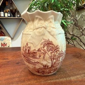 May include: Cream ceramic vase with a ruffled top and bow details. The vase has a brown landscape scene with buildings and palm trees. The vase is on a wooden surface. The vase is approximately 20 cm tall.