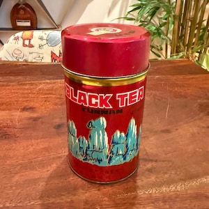 Può includere: Un contenitore cilindrico per tè in metallo rosso con coperchio rimovibile. La parte anteriore presenta la scritta BLACK TEA YUNNAN in caratteri bianchi e neri, accompagnata da un'illustrazione grafica di cime montuose blu e bianche.