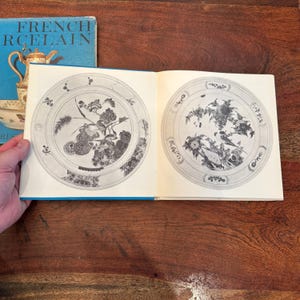 May include: Open book with illustrations of decorative plates. The plates feature floral and bird designs in black and white. The book's cover is blue with the words "FRENCH PORCELAIN" in white.
