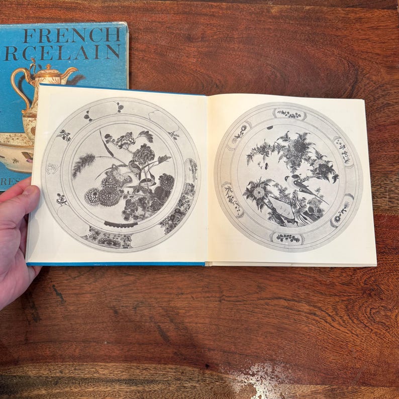 May include: Open book with illustrations of decorative plates. The plates feature floral and bird designs in black and white. The book's cover is blue with the words "FRENCH PORCELAIN" in white.