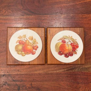 Puede incluir: Dos azulejos de cerámica enmarcados en madera con ilustraciones de frutas. El azulejo de la izquierda presenta una pera, una manzana y bayas verdes. El azulejo de la derecha presenta un melocotón y cerezas.