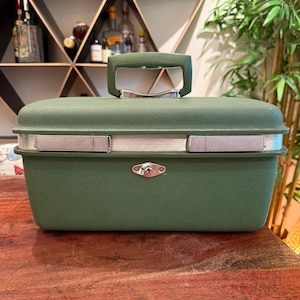 May include: A rectangular olive green hard-shell carrying case with a textured finish. The case features a matching green top handle, a silver-toned metal band encircling the middle, and a central silver-toned metal locking mechanism with a keyhole.
