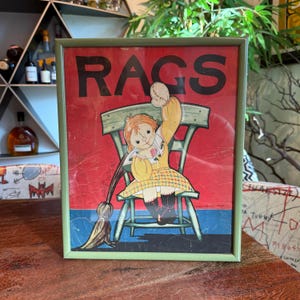 May include: Framed vintage print with the word "RAGS" in large black letters. The artwork features a doll in a yellow dress sitting on a chair, with a broom. The background is red, and the frame is green.